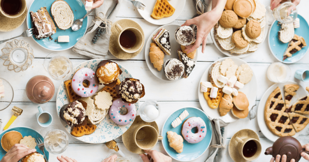 A table filled with assorted desserts, including donuts, ice cream, waffles, cookies, pastries, and cups of coffee and tea—perfect to curb late night cravings—with several hands reaching for treats.