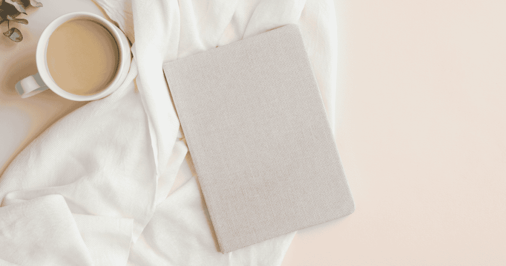 A beige notebook rests on a soft white fabric next to a cup of coffee with milk, creating a calm, cozy scene on a light-colored surface—perfect for jotting down affirmations for healthy eating.