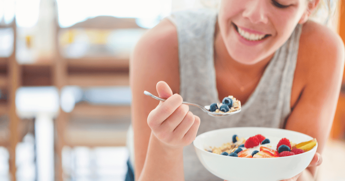 A woman smiles while holding a bowl of oatmeal topped with fresh fruit, including blueberries, raspberries, and strawberries, nourishing your thyroid as she lifts a spoonful to eat.